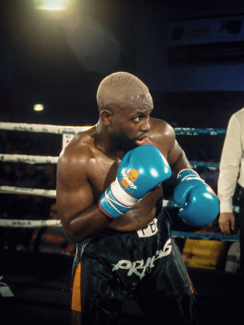 Muskulöser Boxer mit blonden Haaren, schweißbedeckt, in blauen PRIMOS-Boxhandschuhen und schwarzer Shorts, in Kampfpose im Ring.